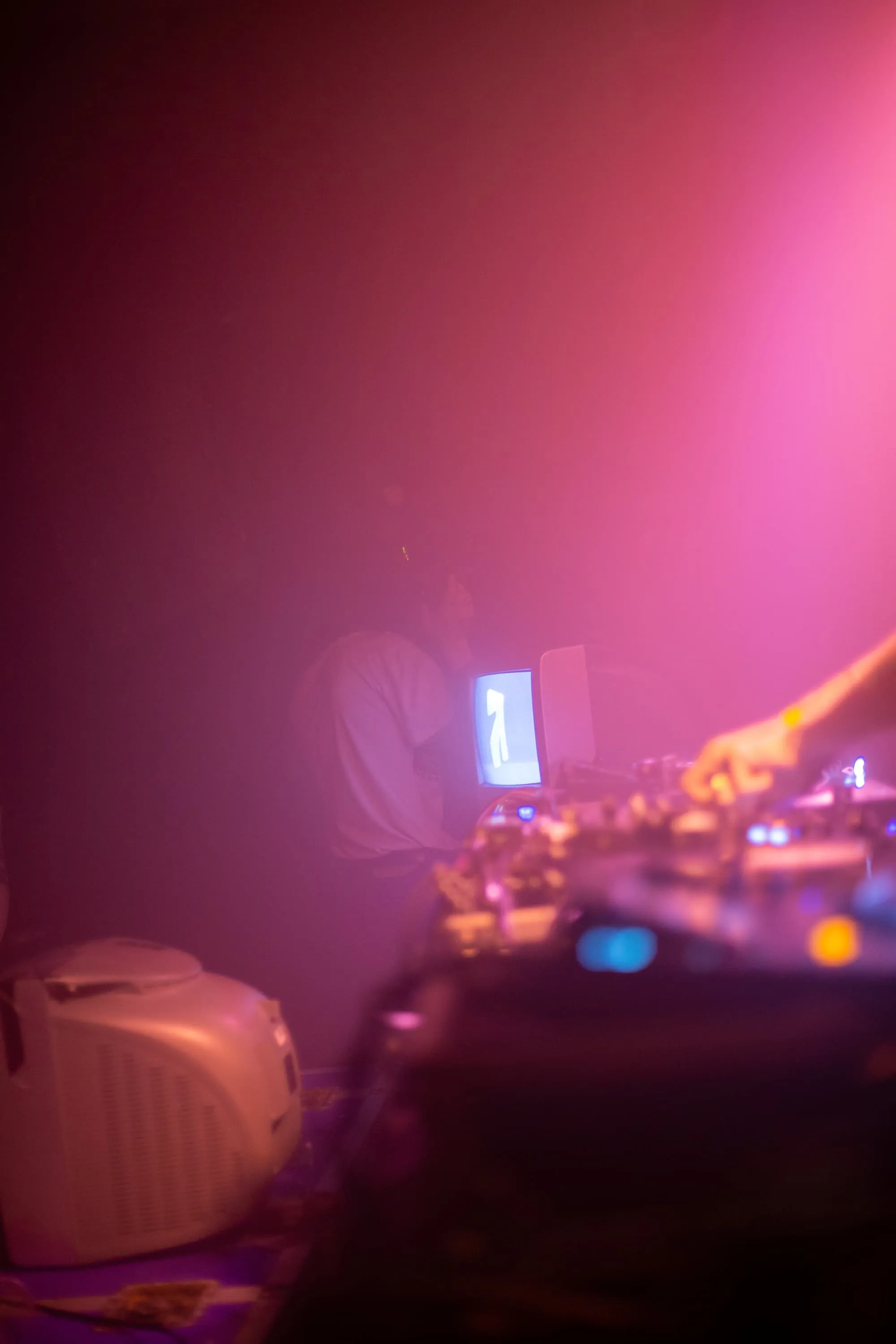 DJ Equiptment in foreground with CRT TV showing 3D metallic logo in the background. A pink hue fills the image from the top right