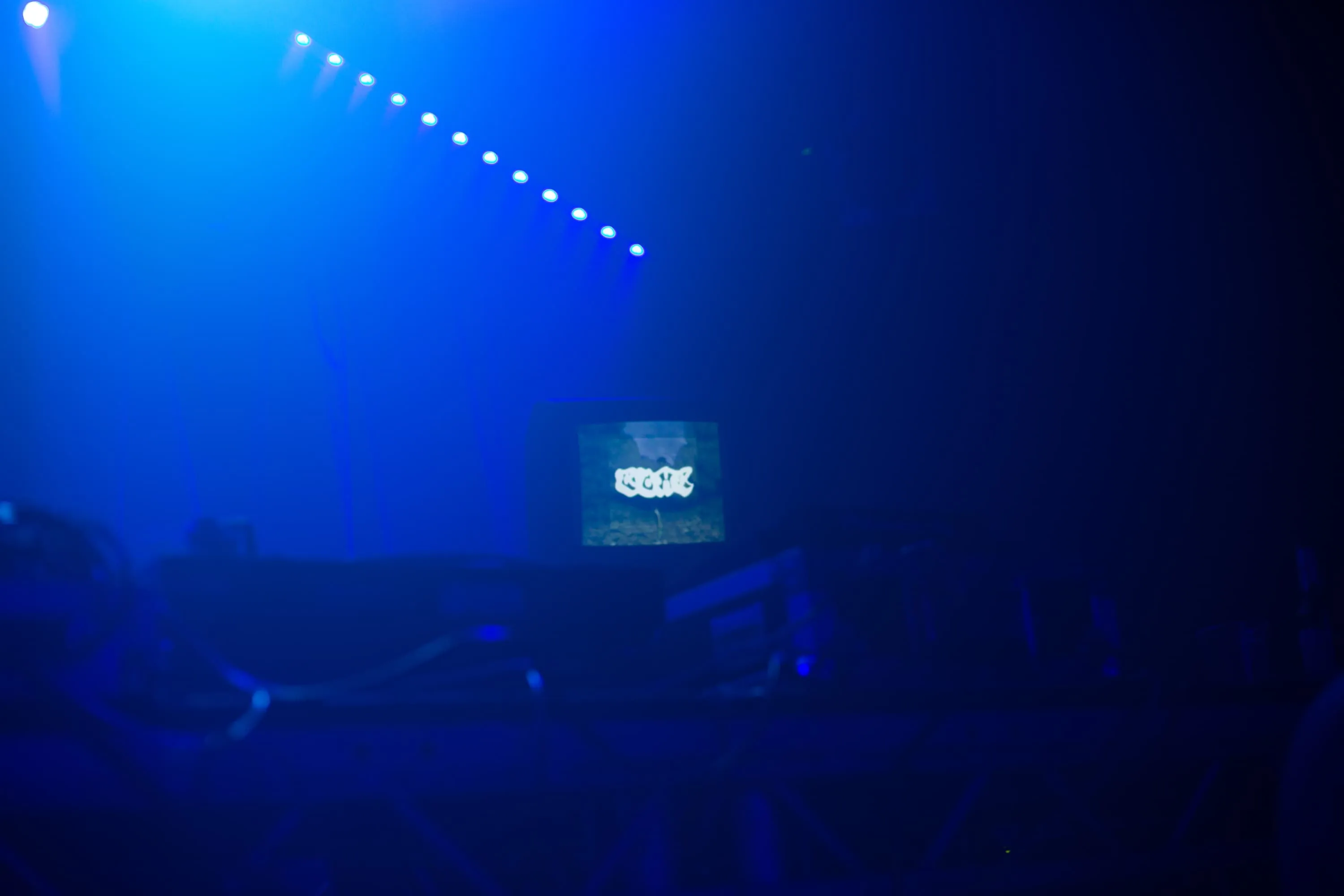 Club space is lit in blue with table holding CDJs in foreground and CRT TV with visuals in the background
