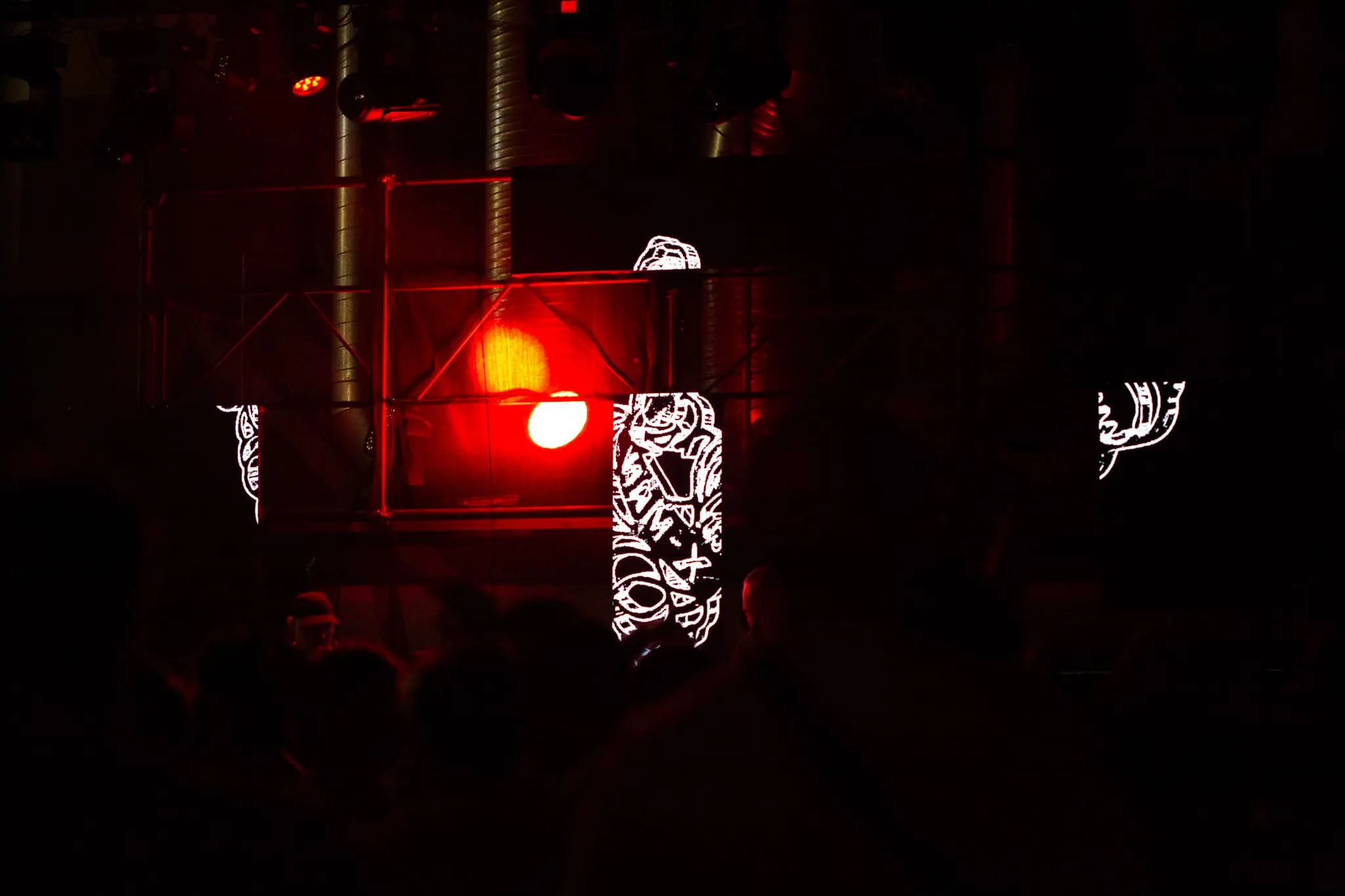 A red light from behind scaffolding lights the scene in a dim pink light. In the foreground you can just make out the crowd and in front of them ia stage with two DJS and 4 LED panels behind them showing a sketchy figure in white.