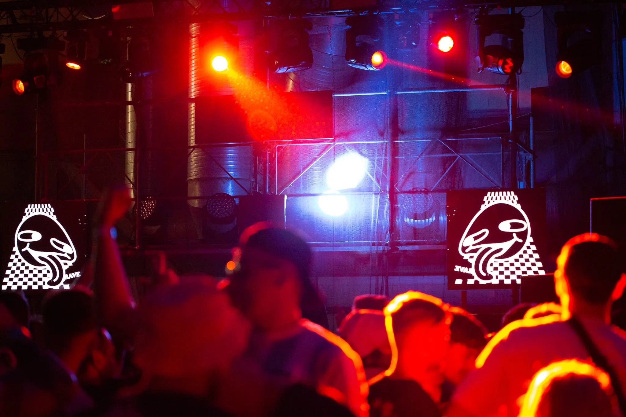Red lights alluminate members of the crowd, in front of them you can see two square LED panels attached to scaffolding with a white graphic on them with the word rave and a face sticking its tongue out.