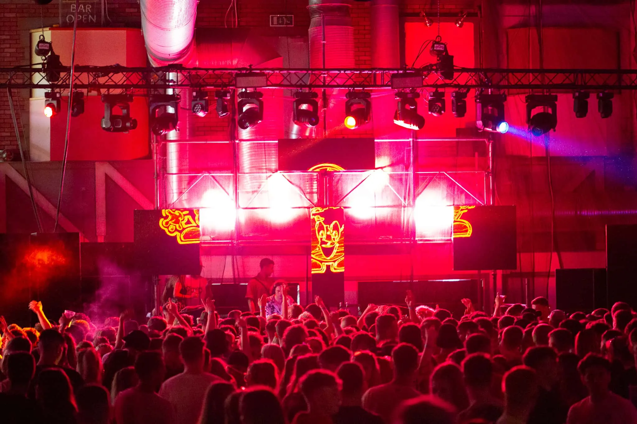 A large crowd in the foreground standing in front of Mall Grab who is DJing with LED panels attached to scaffolding in the background. The panels show a happy looking red character split mapped across all 4 panels. Red light from behind the scaffolding fills the space with a pink hue.