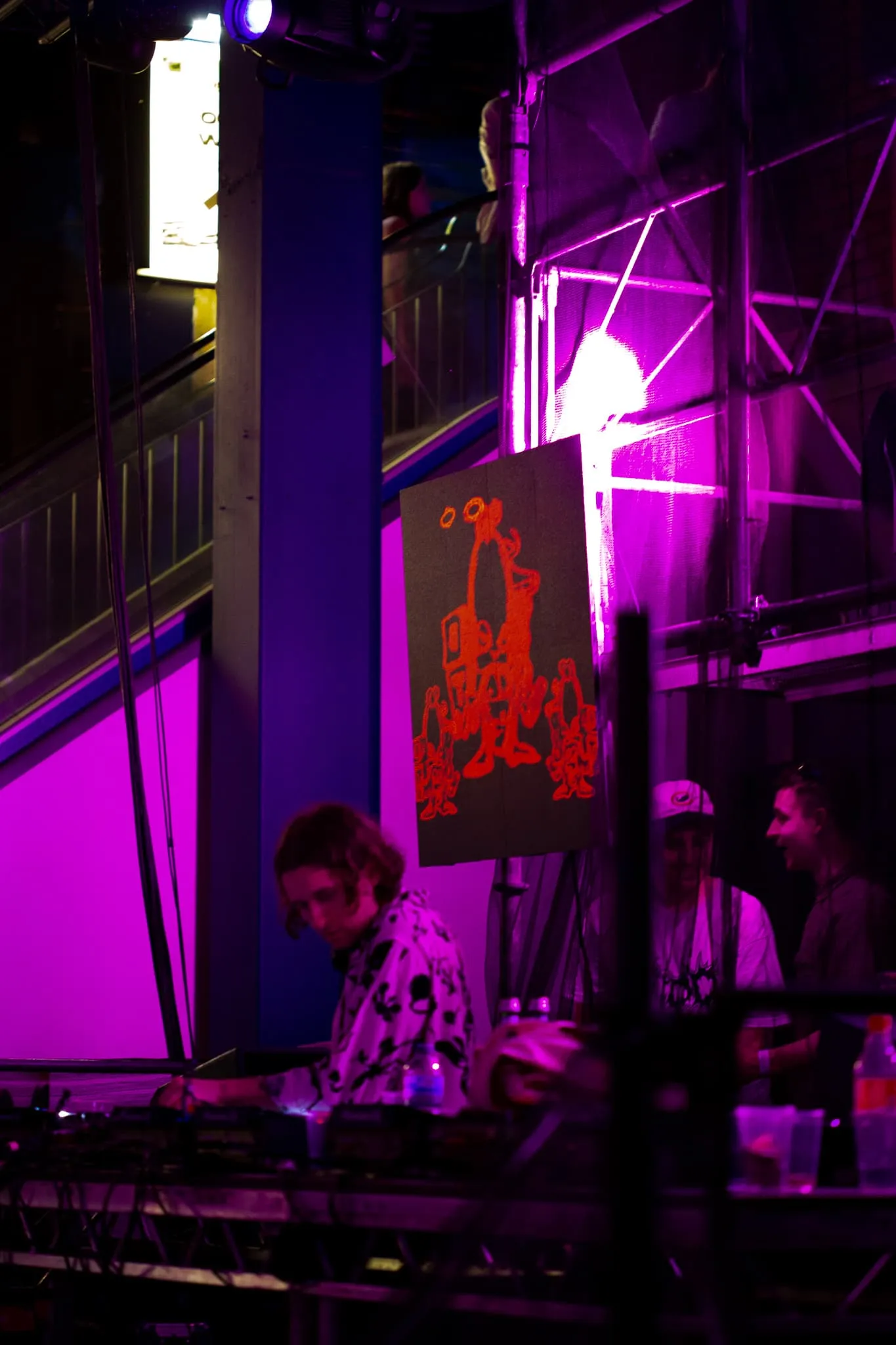 Mall grab DJing in front of scaffolding with square LED panel attached showing naughty looking character holding gun with smoke coming from it (bad tunes logo) in red.
