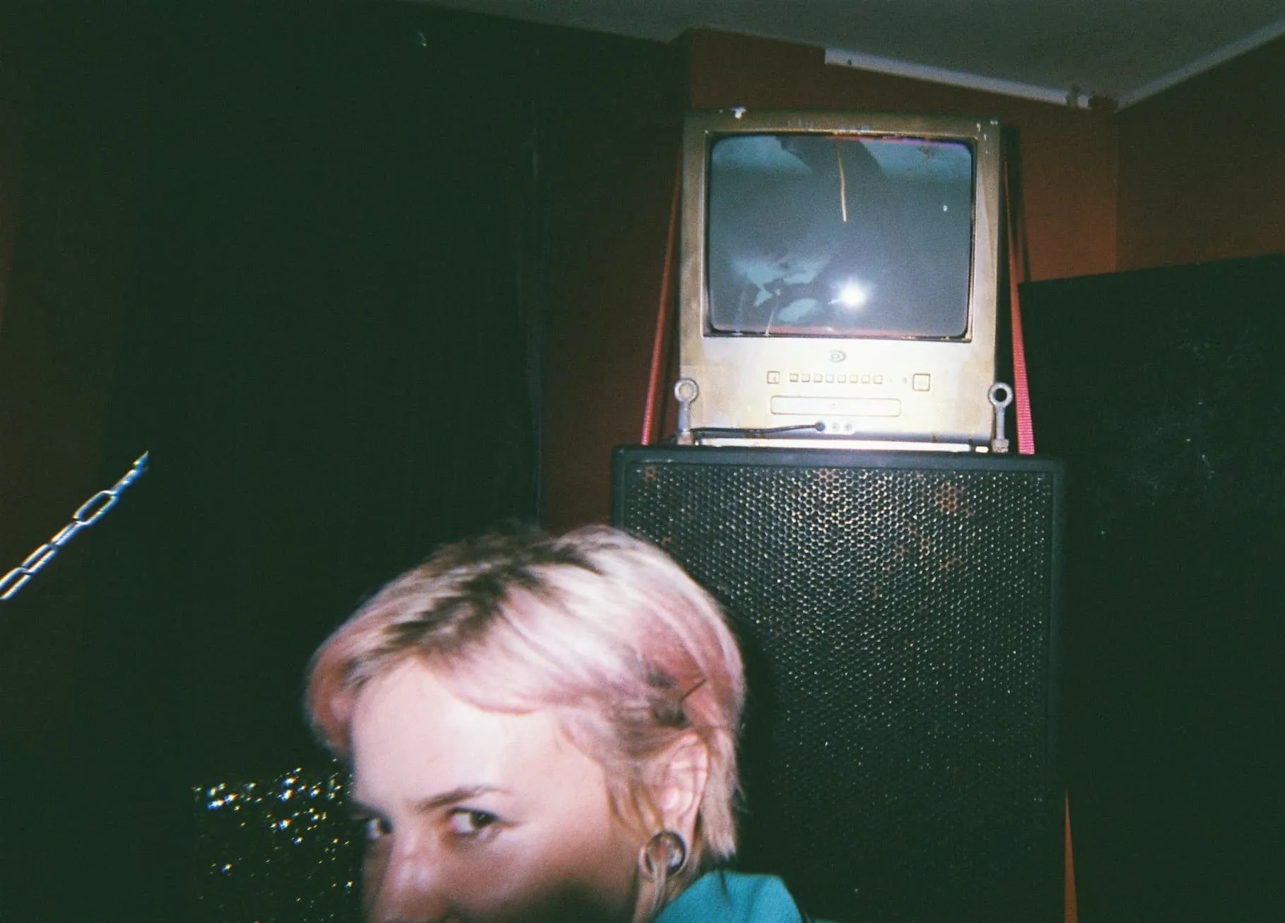 a white woman with pink hear looks smiles at the camera standing in front of a stack of speakers with a golden spray painted CRT TV standing on top of them.