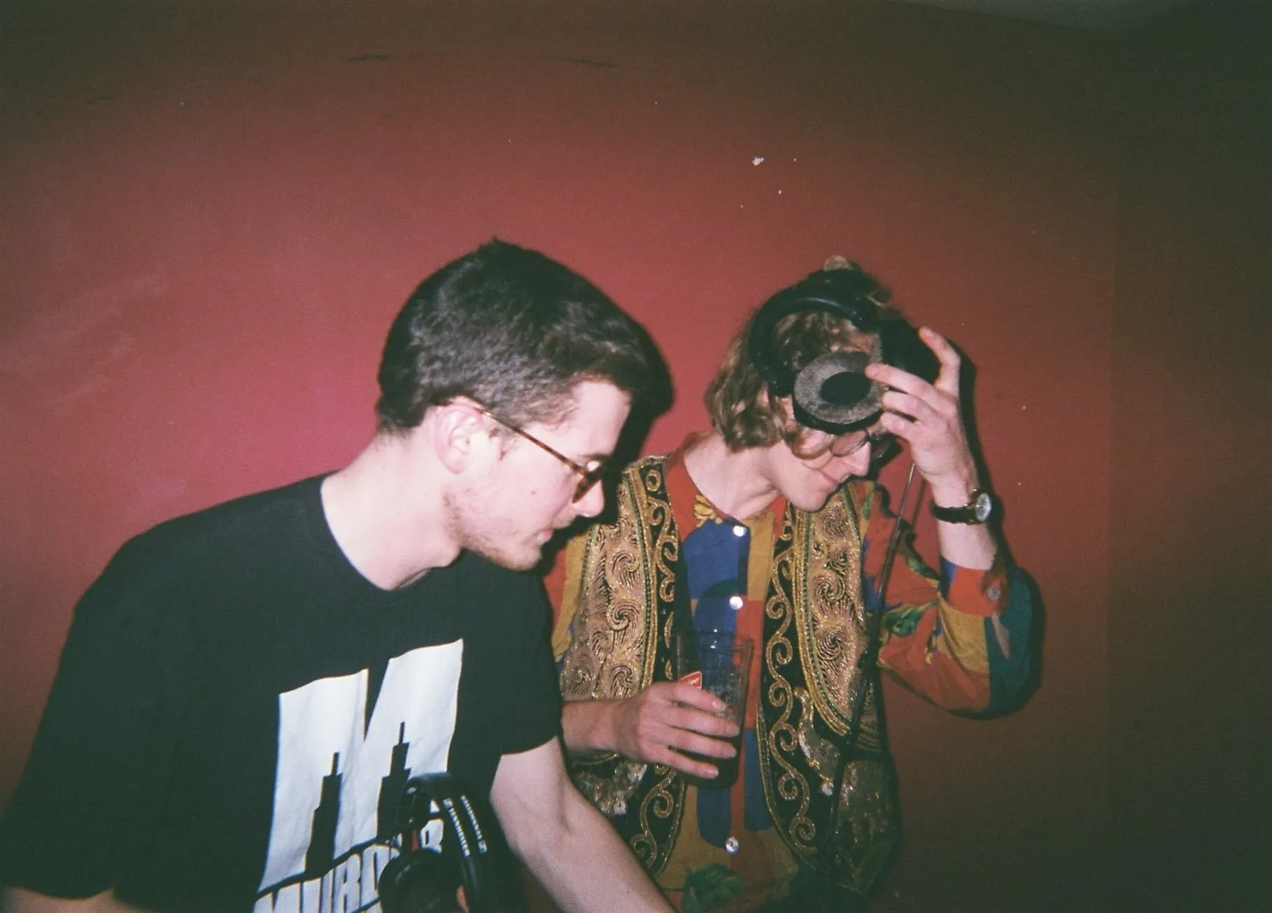 Two white men hold headphones whilst djing, one wearing a black t shirt and the other wearing a glittery golden waistcoat is in the action of removing the headphones from his head. They both stand in front of a red wall.