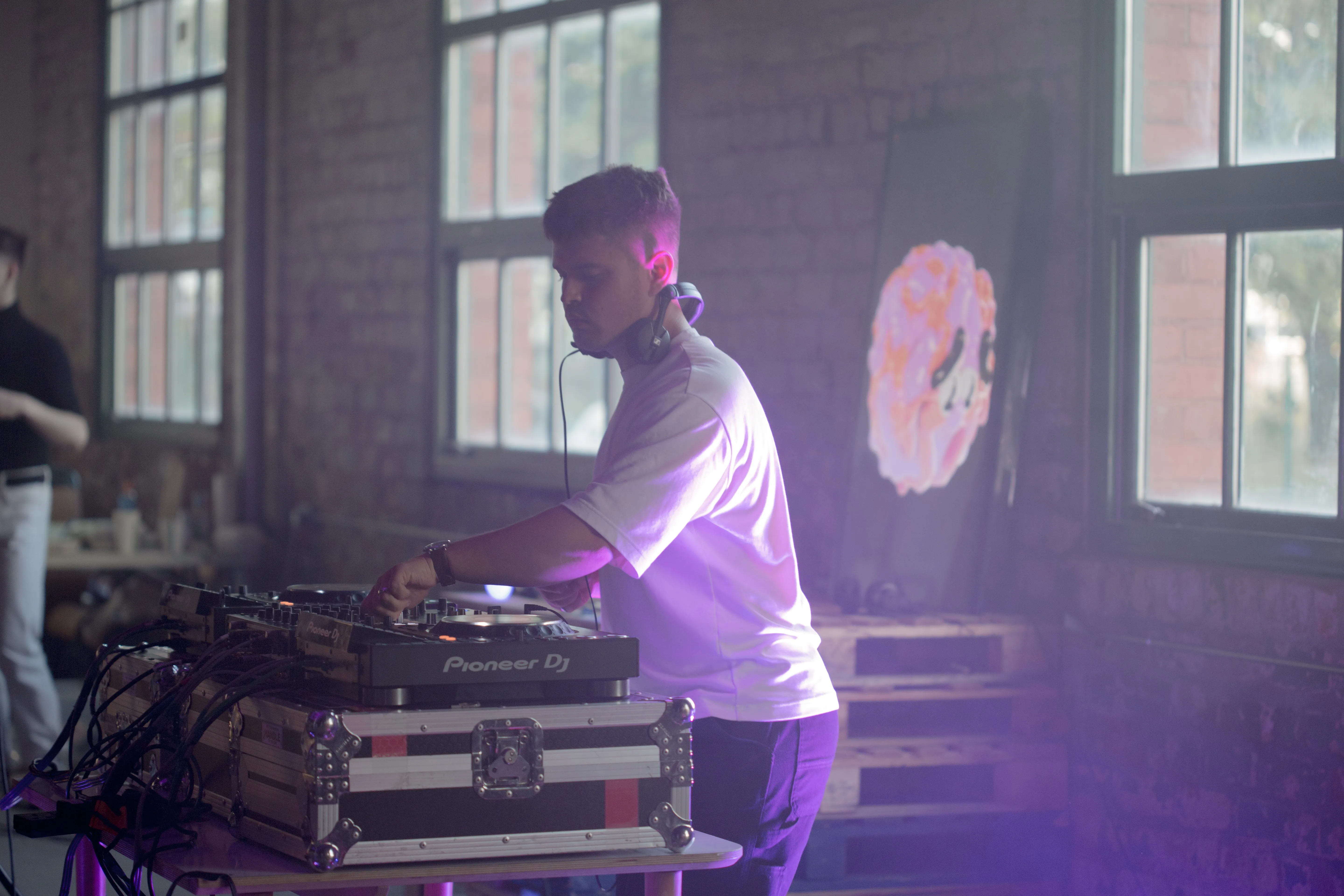 A man DJs light by purple light and sunlight from the windows behind him, in the background a TV shows a 3D heads looking towards the DJ.