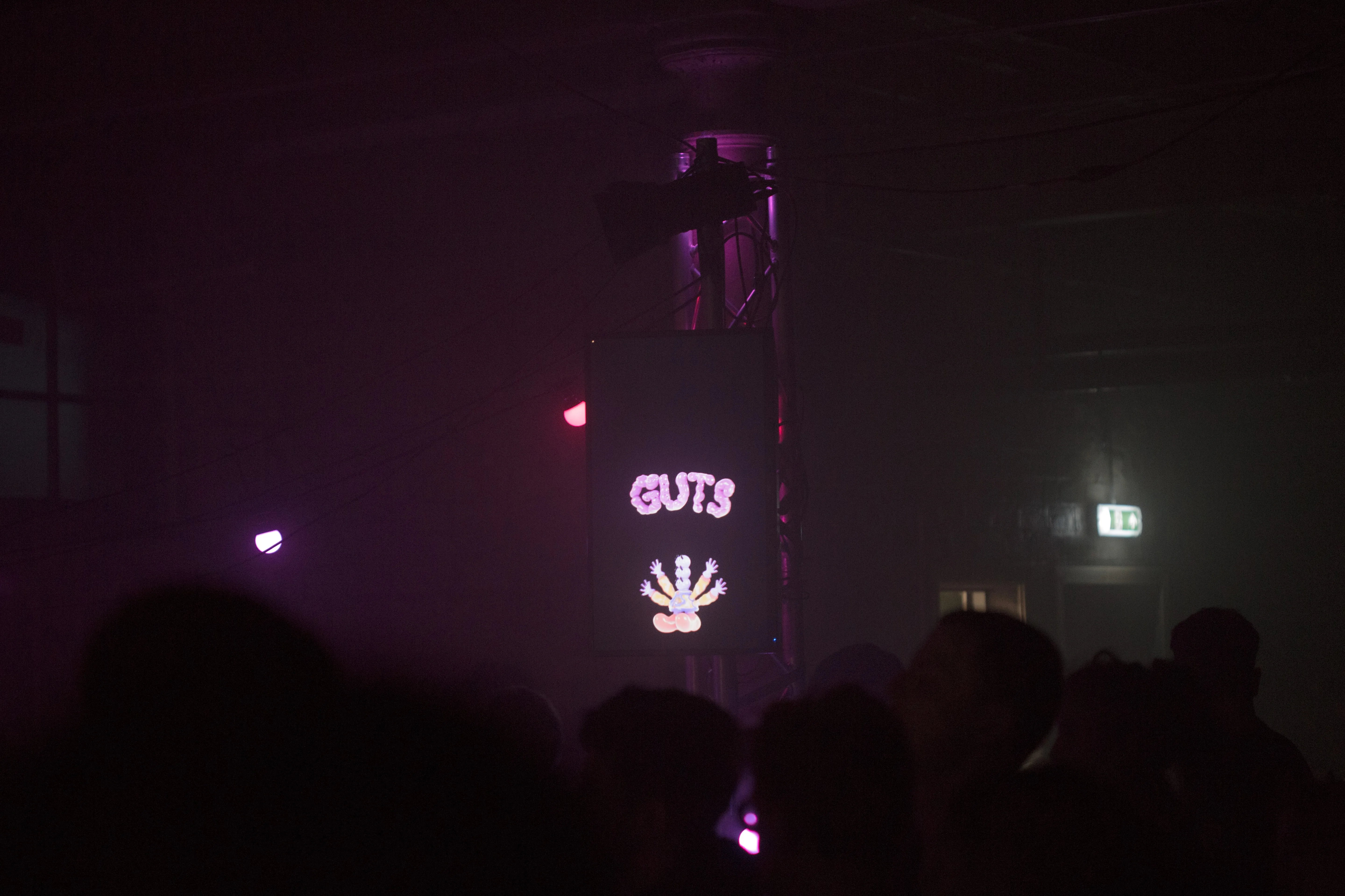 A TV showing the words GUTs in wobbly 3D font and a 3D character sits above a crowd of people's heads.