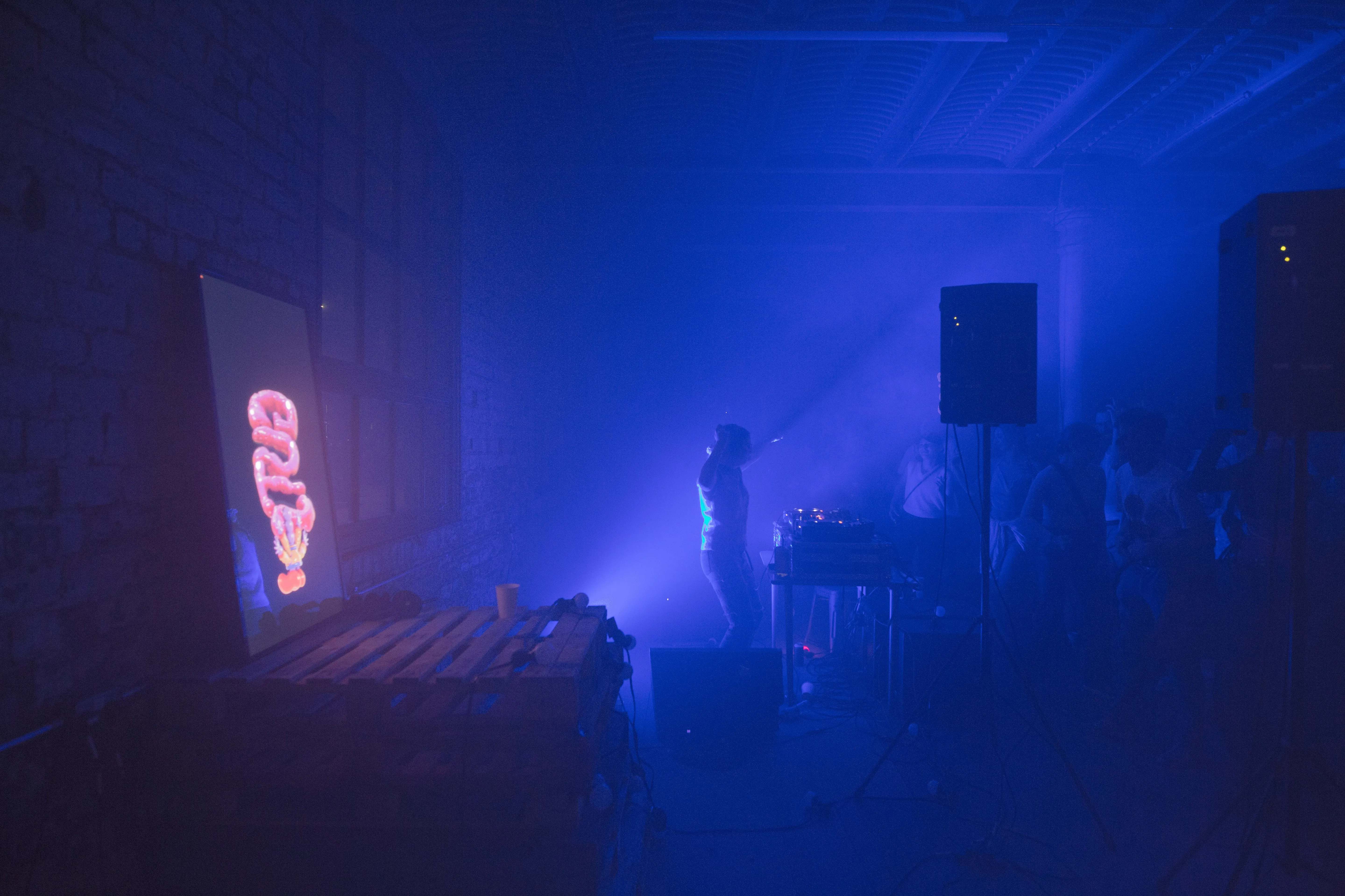 A DJs Jumps hands in the air as the crowd dance barely visibly, in the left of a the photo a TV shines with the 3D wobbly font reading GUTS