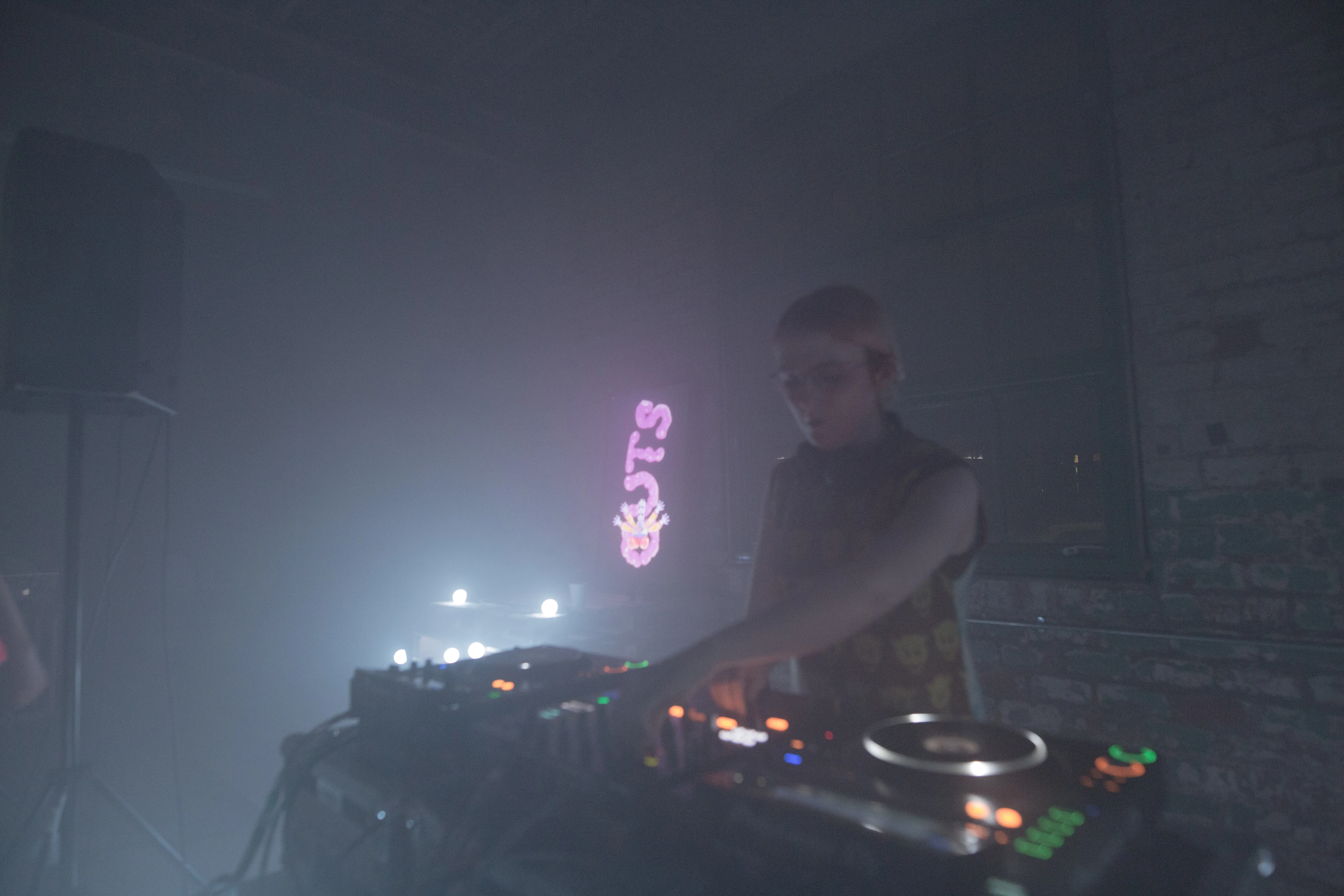 A woman DJs in a dimly lit industrial space with the light from a TV showing GUTS in a wobbly 3D font glowing behind her