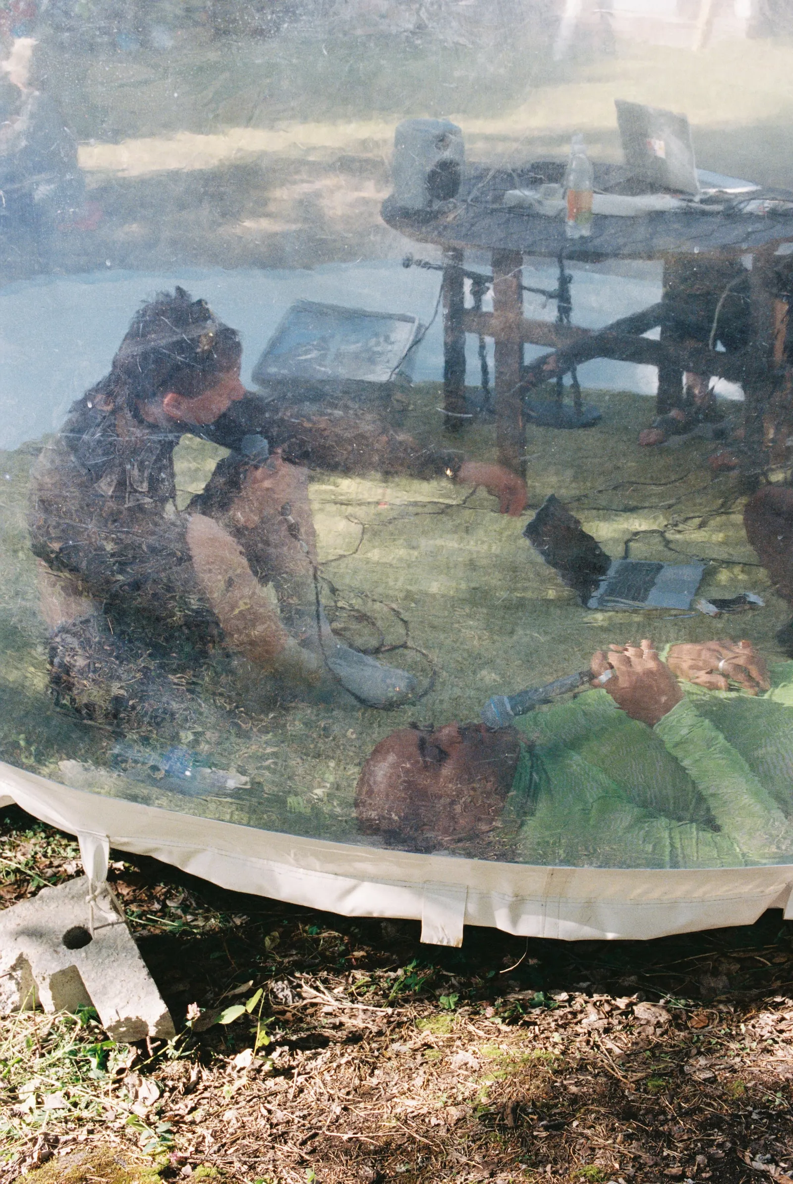 Two people laying down inside a transprent bubble talk on handgeld mics, blurred by the plastic of the bubble