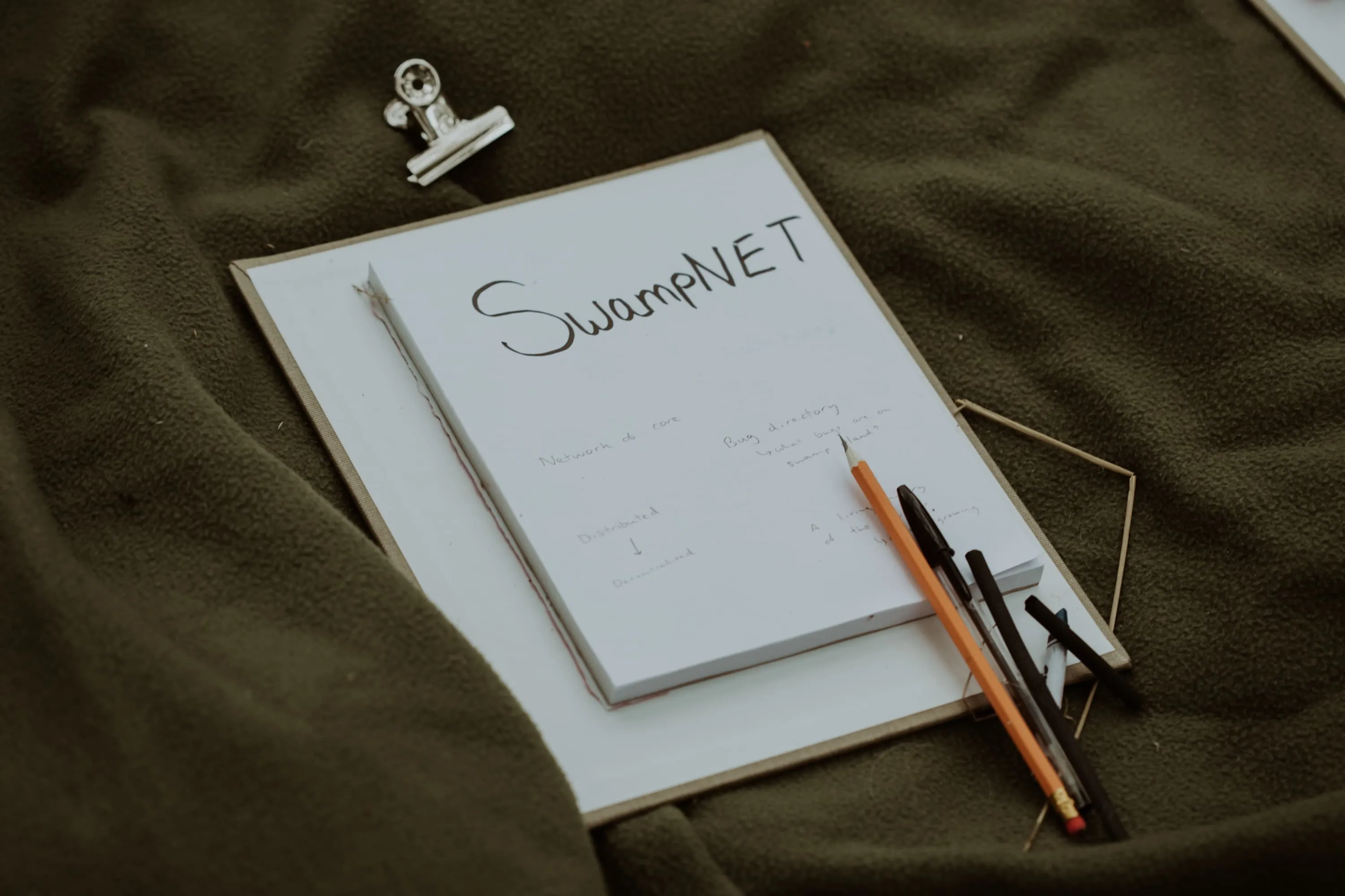 SwampNet is written on a notepad that is held to a clipboard with some pens and pencils next to it. all of this is lying on a nice green blanket.