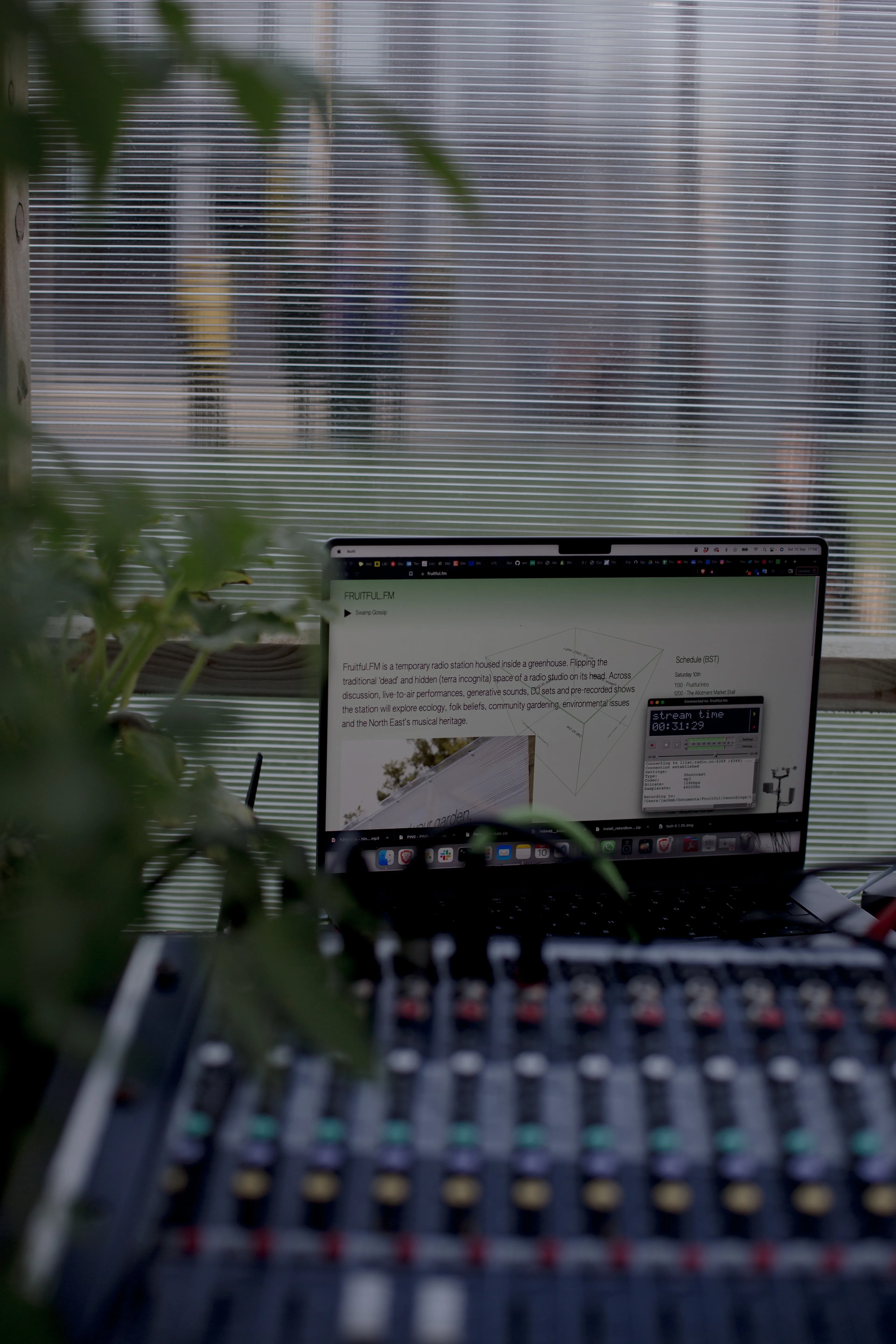 A latop with sits amongst plants and a mixing desk inside a greenhouse