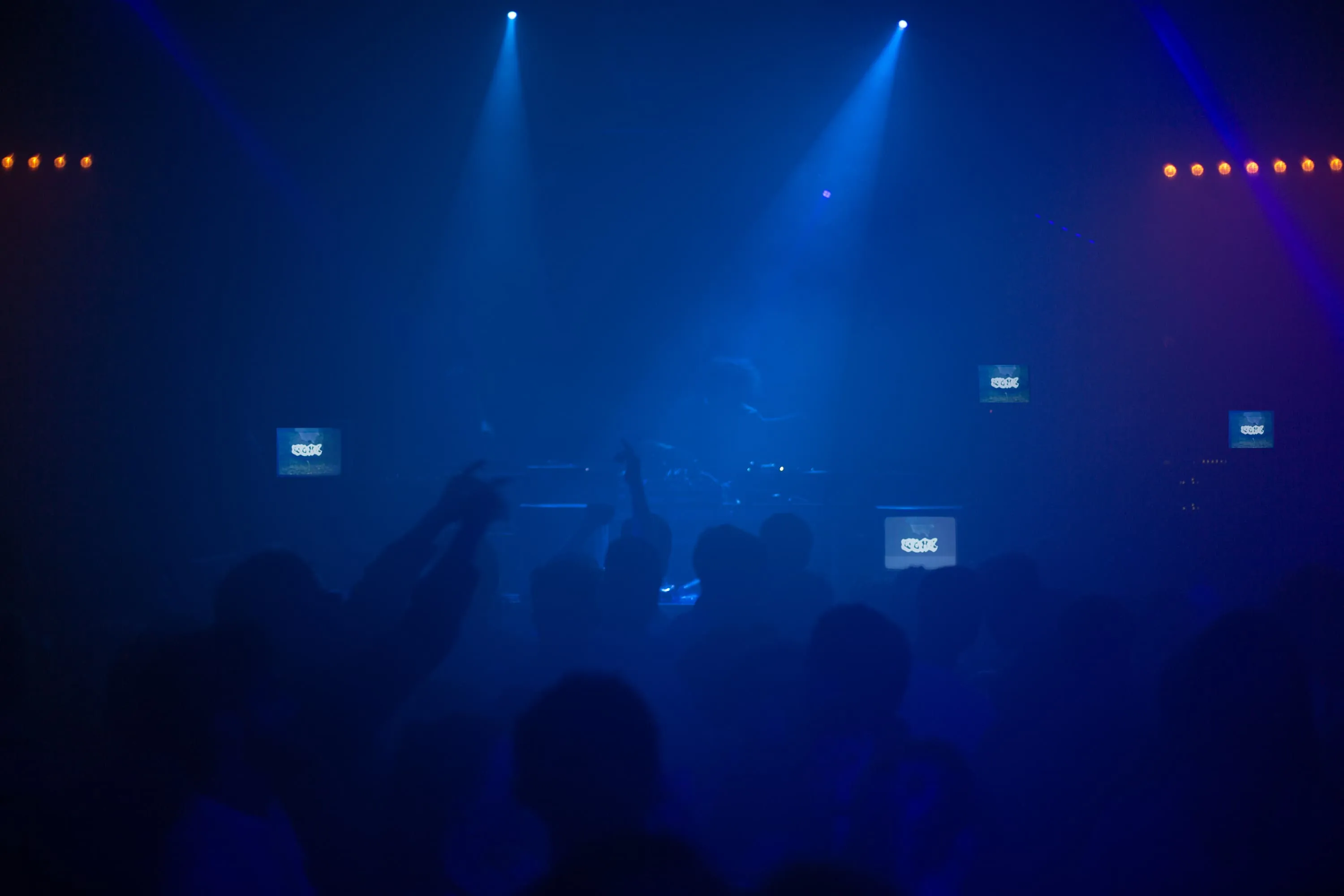 A night club with crowd in foreground having a great time with their arms raised, 4 CRT TVs show visuals spread across the stage and Interplanatary Criminal DJs in the centre of the stage with a beam of light on him