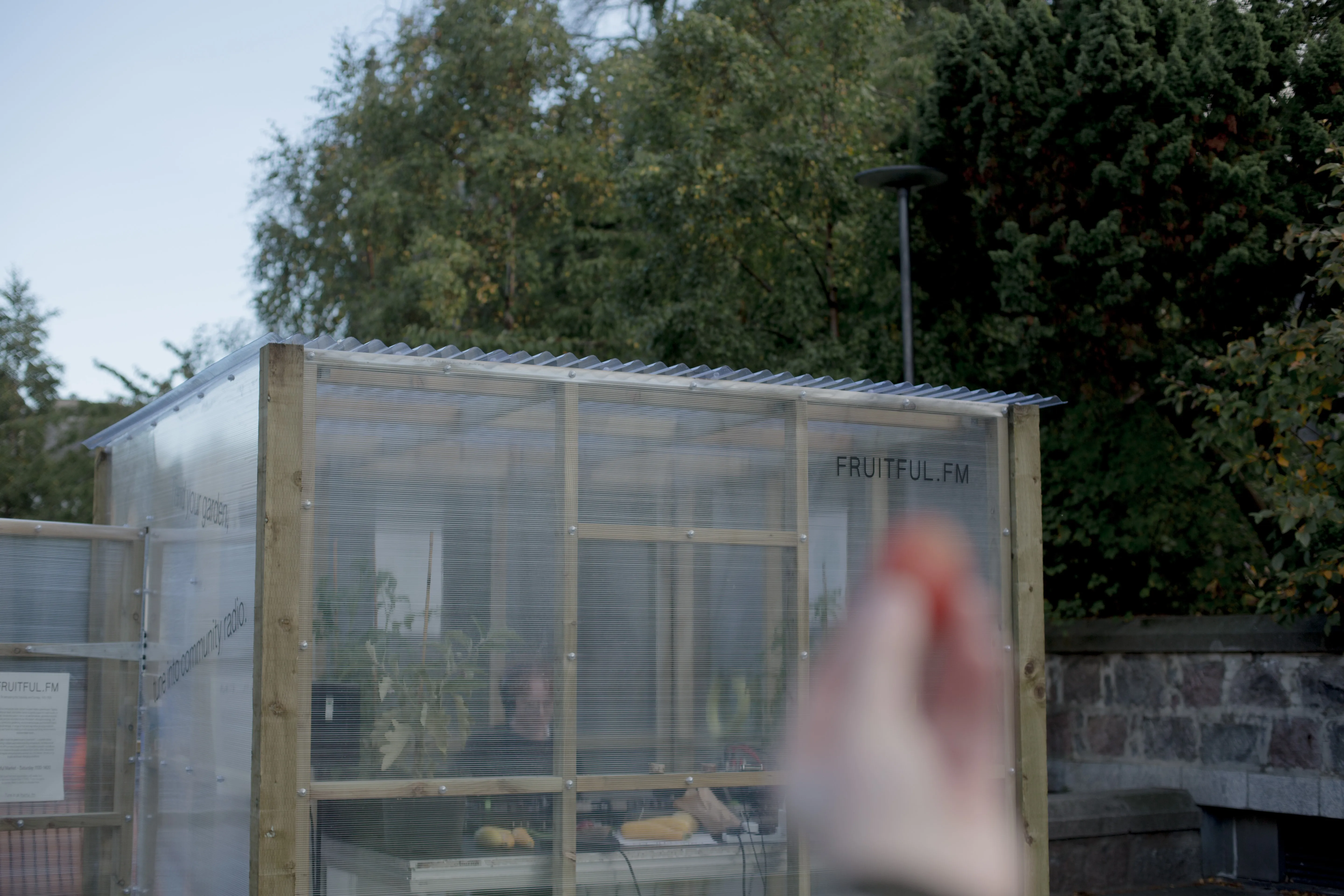 A blurred plum is held in front of a greenhouse with vinyl reading Fruitful.FM visible