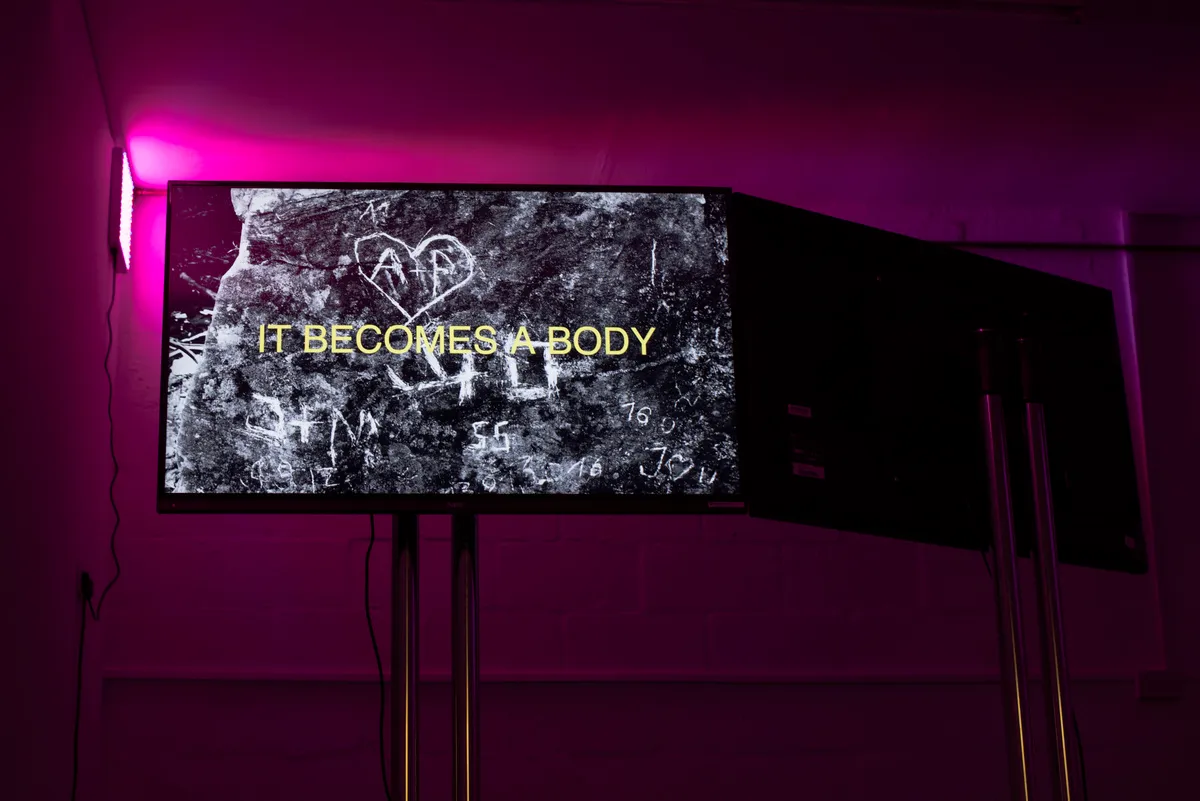 Pink light at the top of the image fills with basement space with a nice pinky glow. A tv showing the words 'It becomes a body' are shown a tv on a stand.