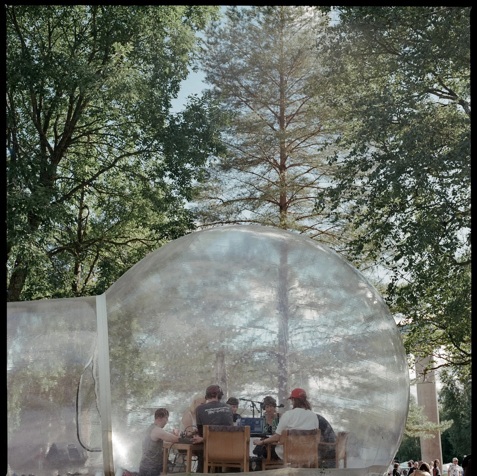 A group of people sit around a table with mics inside a transarent bubble in amongst green trees