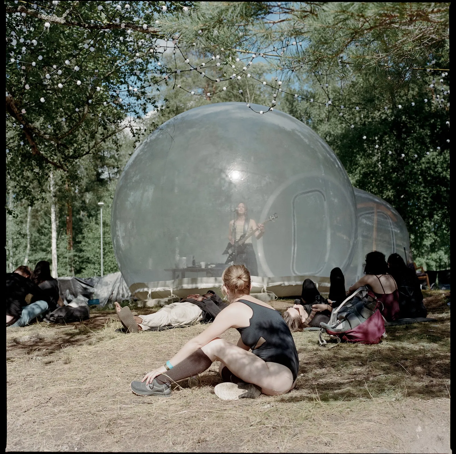 A person sits arm streched out in a black bathing suit in front of a transprent bubble with a person playing a guitar inside.