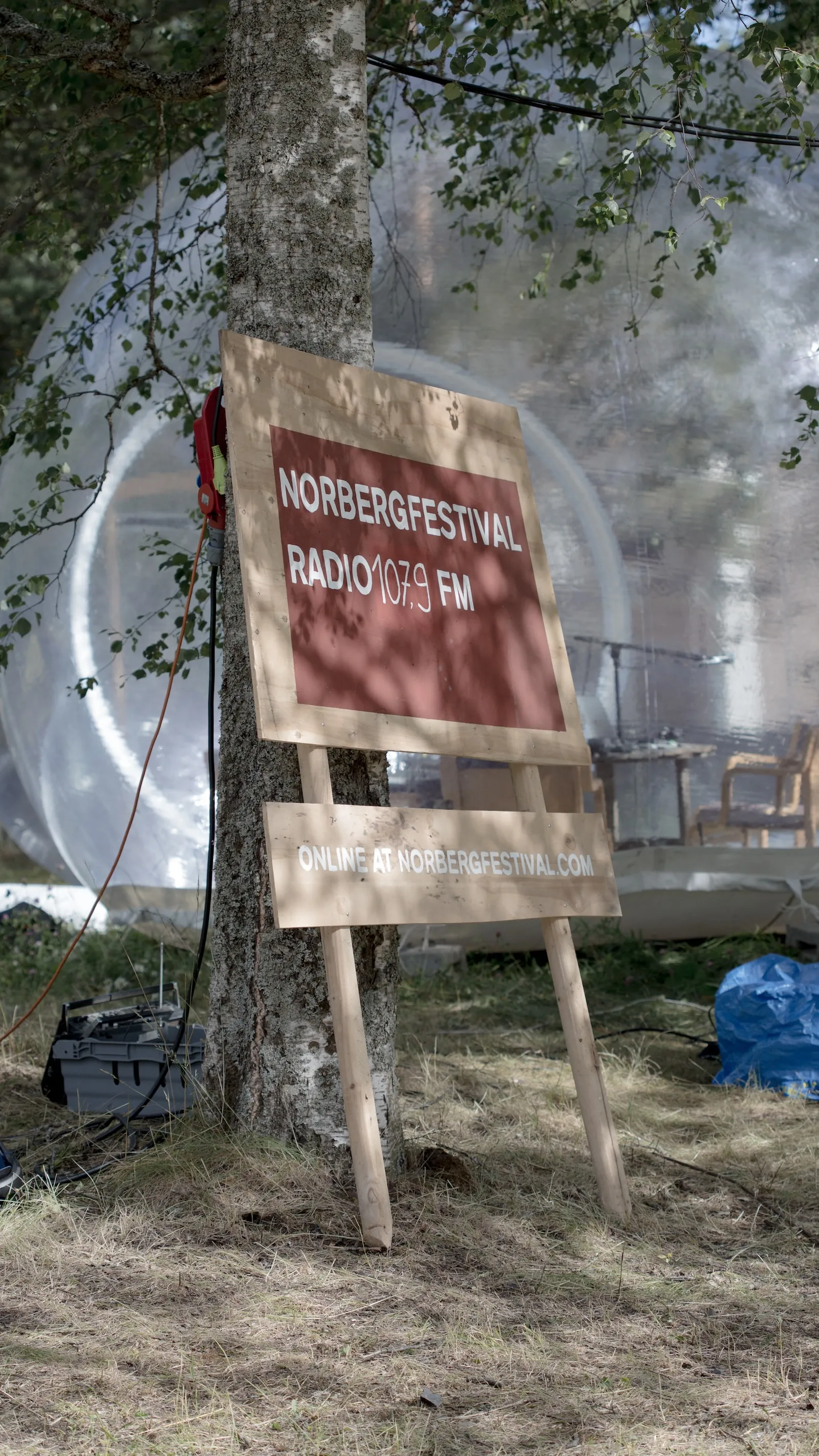 A wooden signs propped up against a tree reads Norbergfestival Radio 107.9 FM