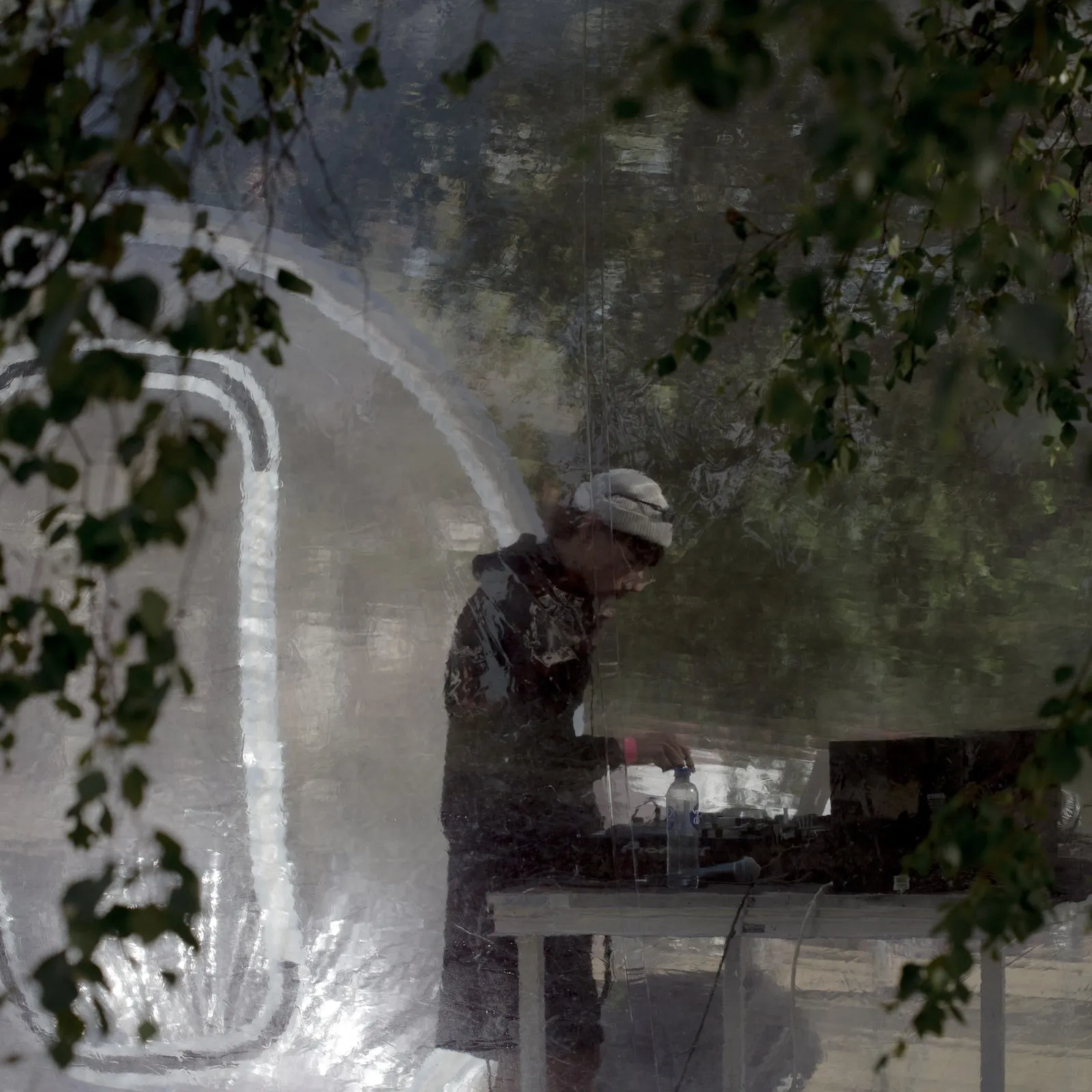 A person DJs inside a transprent bubble blurred by the plastic of the bubble and surrounding green leaves