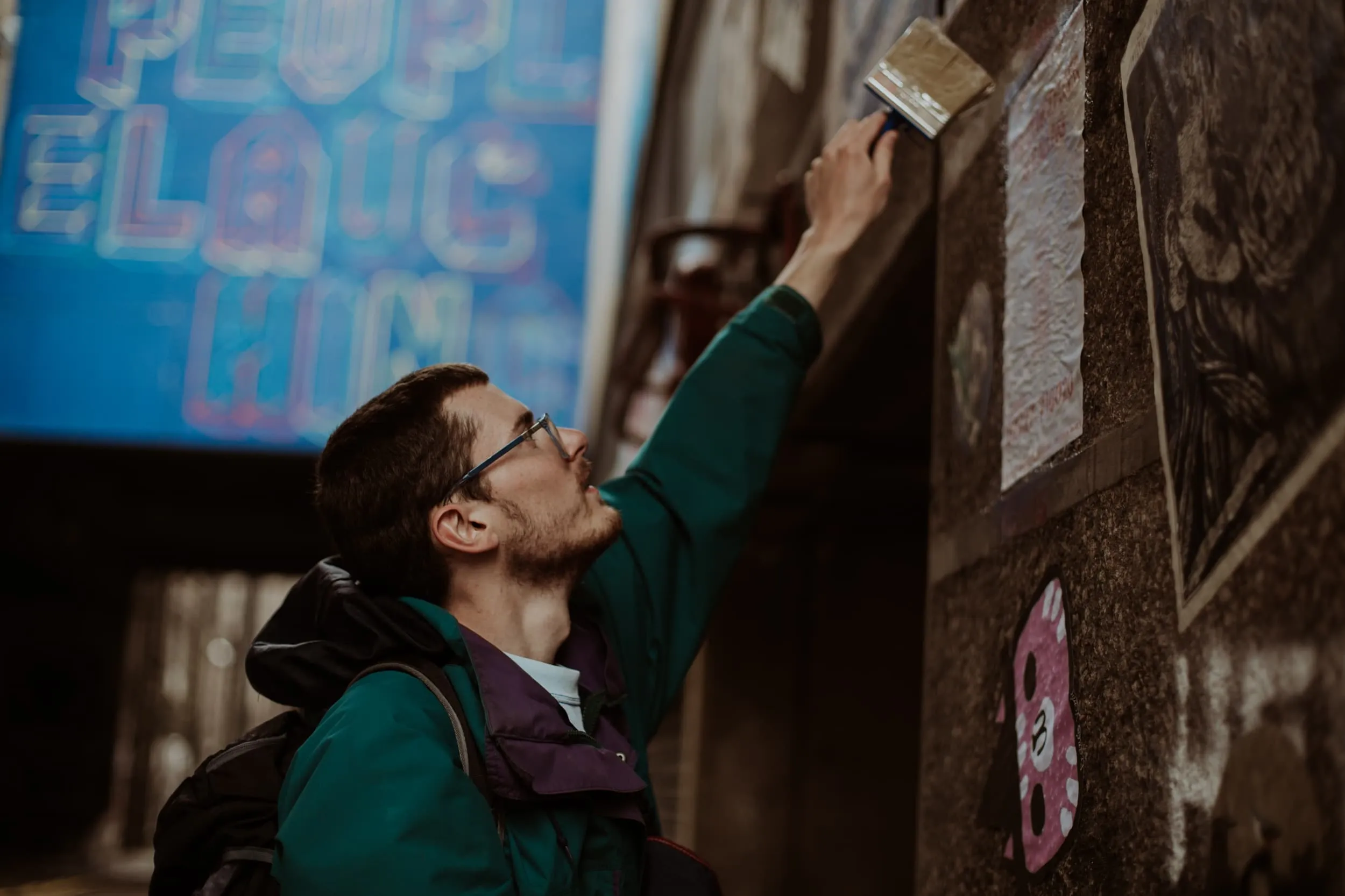 Man with wierd and large paint brush and green jacket pasting up poster on granite wall with bright blue mural blurred in the background
