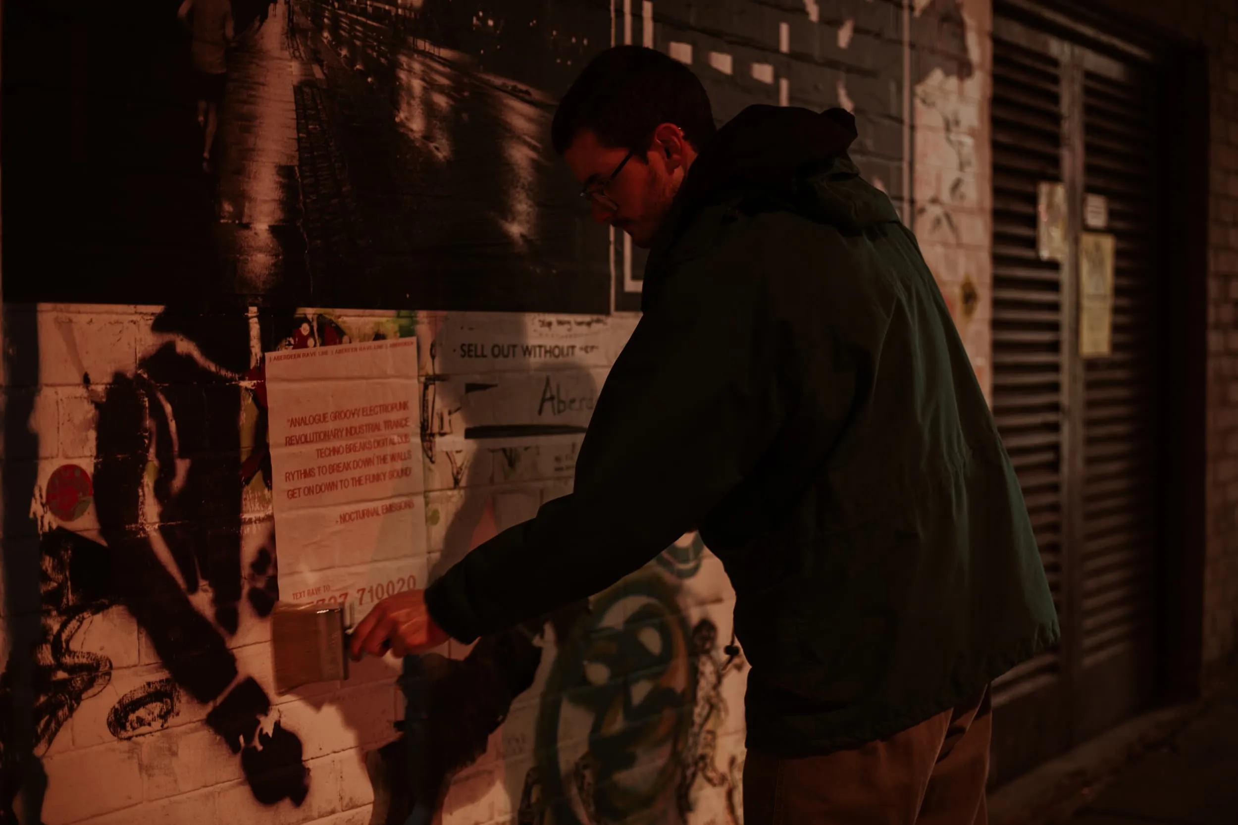 Man with wierd and large paint brush and green jacket pasting up poster on white brick wall